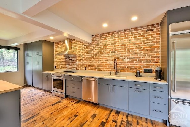 kitchen with wall chimney range hood, sink, light hardwood / wood-style flooring, stainless steel appliances, and brick wall