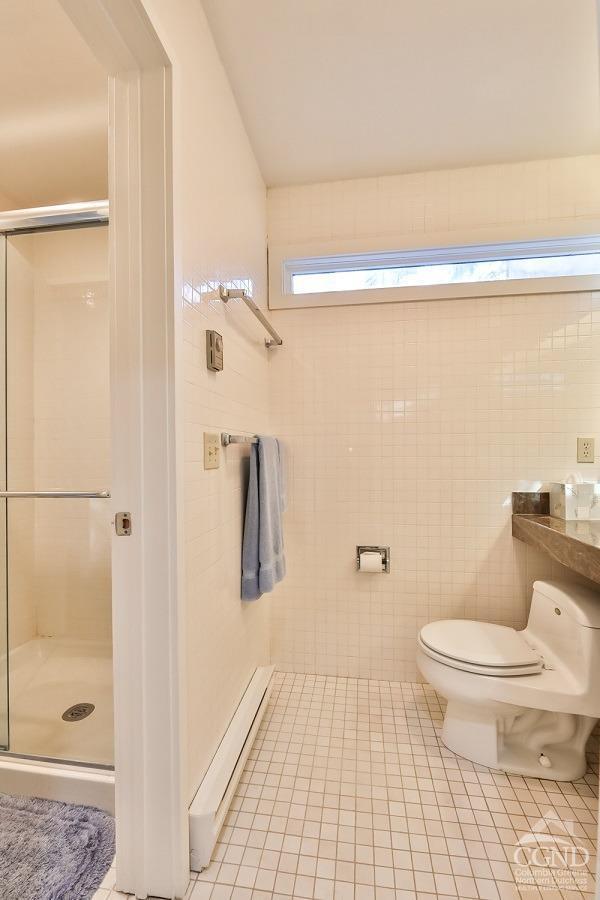 bathroom with tile patterned flooring, a baseboard heating unit, toilet, a shower with shower door, and tile walls