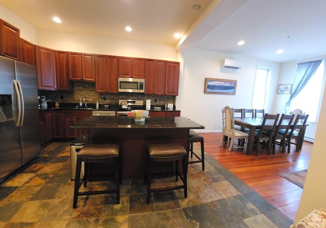 kitchen featuring dark countertops, backsplash, appliances with stainless steel finishes, an AC wall unit, and a kitchen bar