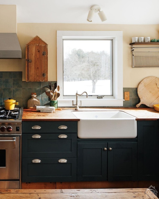 kitchen featuring stainless steel stove, butcher block counters, a sink, backsplash, and wall chimney exhaust hood