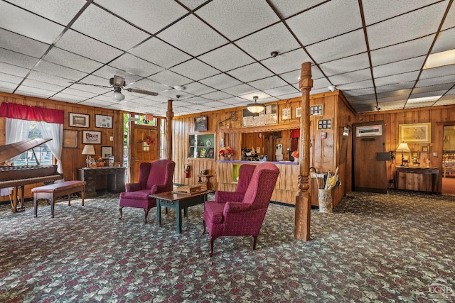 living room with a paneled ceiling, wood walls, ceiling fan, and dark carpet