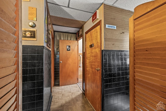 hall featuring a paneled ceiling, wooden walls, tile walls, and parquet flooring