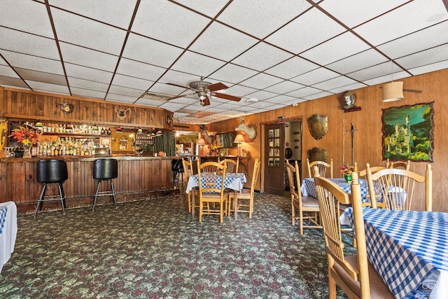 unfurnished dining area with dark carpet, a drop ceiling, ceiling fan, wooden walls, and bar