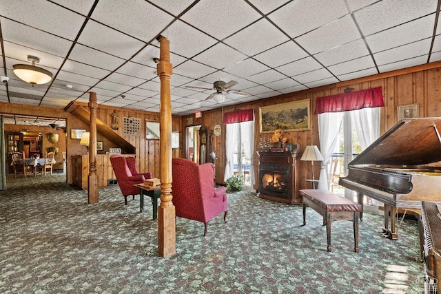 carpeted living room with ceiling fan, a drop ceiling, and wooden walls