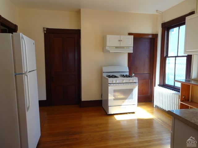 kitchen featuring under cabinet range hood, light wood-type flooring, radiator heating unit, white appliances, and white cabinets