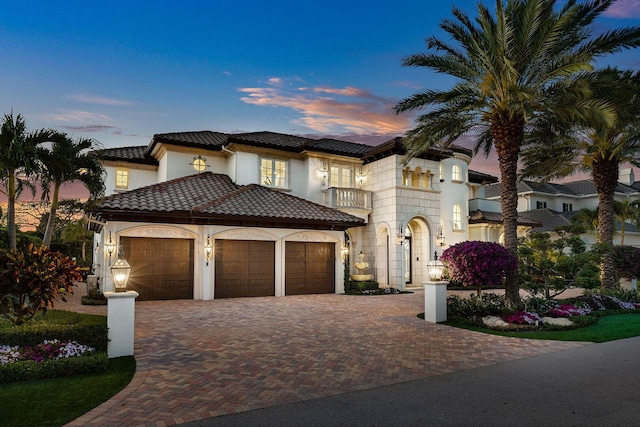 mediterranean / spanish home with stucco siding, decorative driveway, a tile roof, a balcony, and a garage