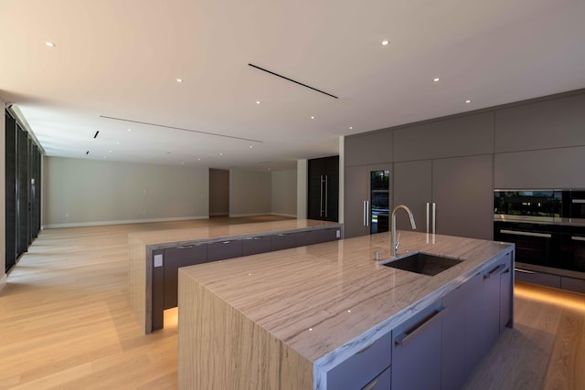 kitchen with sink, light wood-type flooring, a center island with sink, light stone counters, and double wall oven