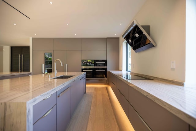 kitchen with an island with sink, ventilation hood, gray cabinets, light wood-type flooring, and light stone counters