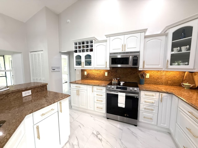 kitchen with glass insert cabinets, dark stone countertops, appliances with stainless steel finishes, a towering ceiling, and marble finish floor