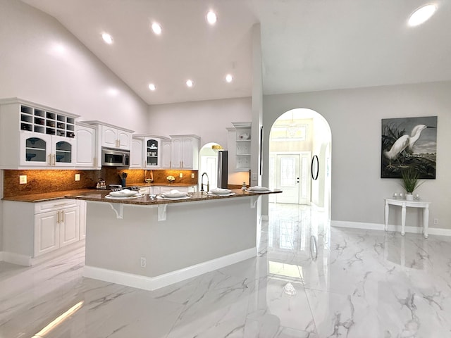 kitchen featuring high vaulted ceiling, open shelves, stainless steel microwave, arched walkways, and glass insert cabinets