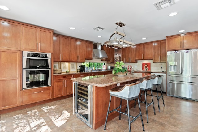 kitchen with an island with sink, stainless steel appliances, wall chimney exhaust hood, hanging light fixtures, and light stone counters