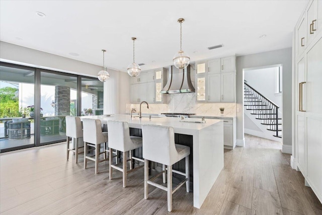 kitchen featuring an island with sink, tasteful backsplash, wall chimney exhaust hood, hanging light fixtures, and light wood-type flooring