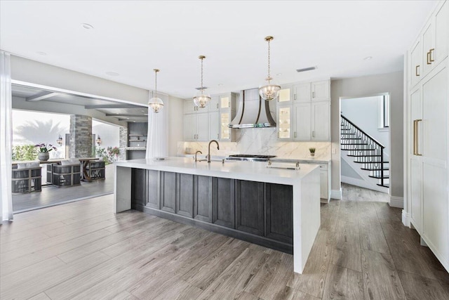 kitchen featuring backsplash, a kitchen island with sink, light hardwood / wood-style floors, wall chimney exhaust hood, and pendant lighting