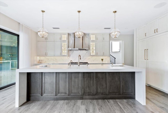 kitchen featuring wall chimney range hood, tasteful backsplash, white cabinets, hanging light fixtures, and light wood-type flooring