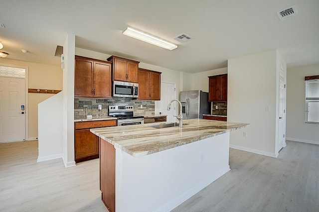 kitchen featuring stainless steel appliances, tasteful backsplash, a center island with sink, sink, and light stone counters