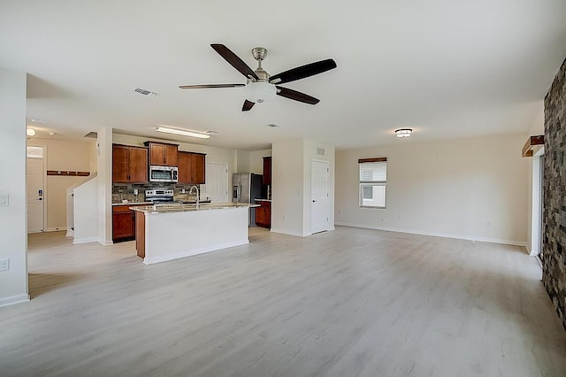 kitchen with a kitchen island with sink, ceiling fan, appliances with stainless steel finishes, backsplash, and light wood-type flooring