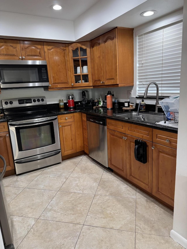 kitchen with appliances with stainless steel finishes, a sink, brown cabinetry, glass insert cabinets, and light tile patterned floors