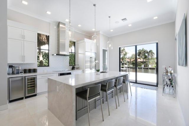 kitchen featuring wall chimney range hood, a kitchen island with sink, stainless steel built in refrigerator, beverage cooler, and backsplash