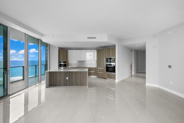 kitchen featuring stainless steel microwave, a kitchen island with sink, white cabinets, a water view, and sink