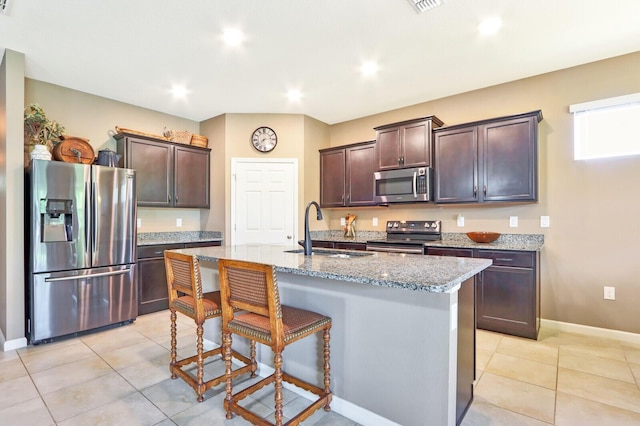 kitchen with light stone countertops, a center island with sink, dark brown cabinets, appliances with stainless steel finishes, and sink