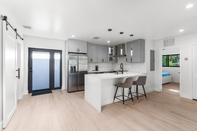 kitchen featuring light hardwood / wood-style floors, kitchen peninsula, a barn door, wall chimney exhaust hood, and stainless steel fridge with ice dispenser