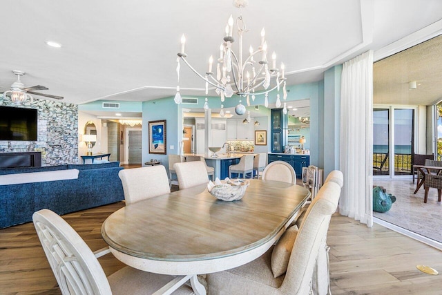 dining area with light hardwood / wood-style floors, ceiling fan with notable chandelier, and a stone fireplace