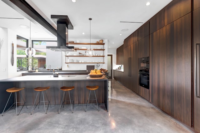 kitchen featuring a kitchen bar, a center island with sink, black double oven, hanging light fixtures, and island exhaust hood