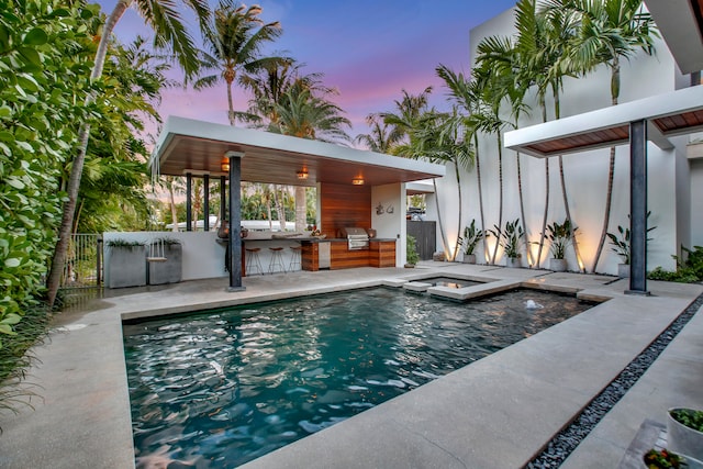 pool at dusk with an outdoor bar, grilling area, exterior kitchen, an in ground hot tub, and a patio