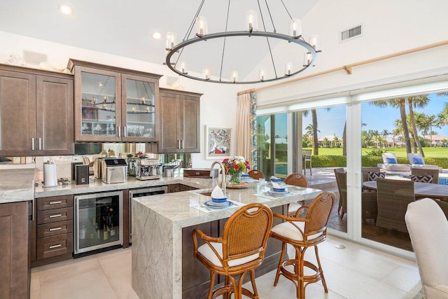 kitchen featuring a notable chandelier, an island with sink, light tile floors, beverage cooler, and light stone counters