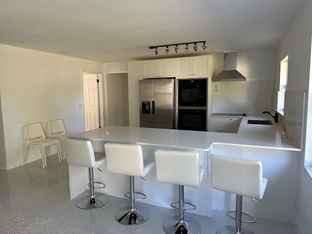 kitchen featuring stainless steel fridge, double oven, wall chimney exhaust hood, a kitchen bar, and white cabinetry