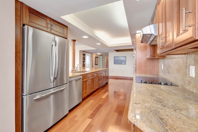 kitchen with ventilation hood, light stone counters, stainless steel appliances, a raised ceiling, and light hardwood / wood-style flooring