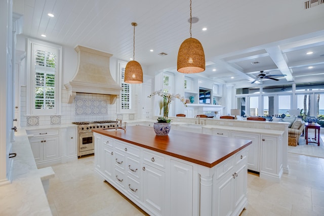 kitchen with coffered ceiling, backsplash, a center island with sink, custom range hood, and double oven range