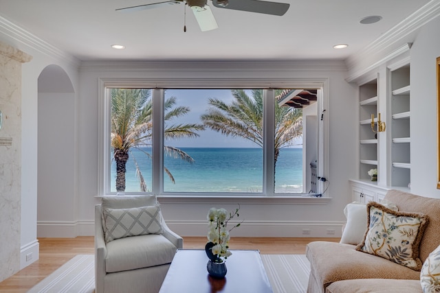 sitting room with ceiling fan, a water view, built in features, light hardwood / wood-style flooring, and crown molding