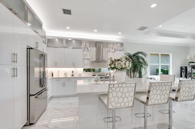 kitchen with high end refrigerator, light tile floors, a breakfast bar area, wall chimney exhaust hood, and white cabinetry