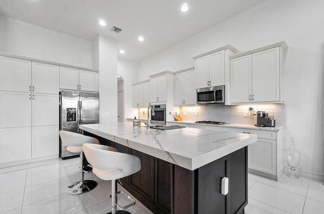 kitchen featuring an island with sink, sink, appliances with stainless steel finishes, light tile flooring, and white cabinetry