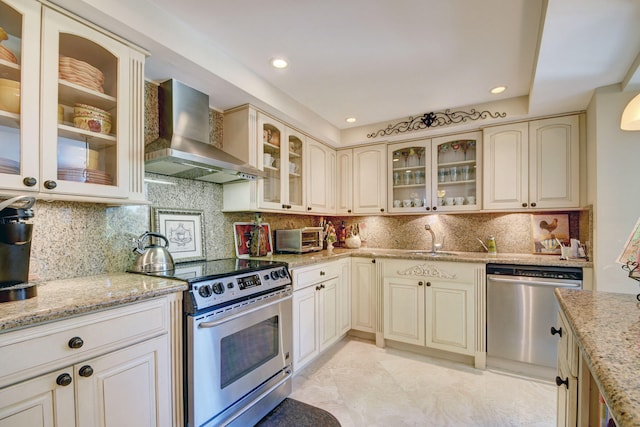 kitchen featuring light stone counters, decorative backsplash, wall chimney exhaust hood, appliances with stainless steel finishes, and cream cabinetry