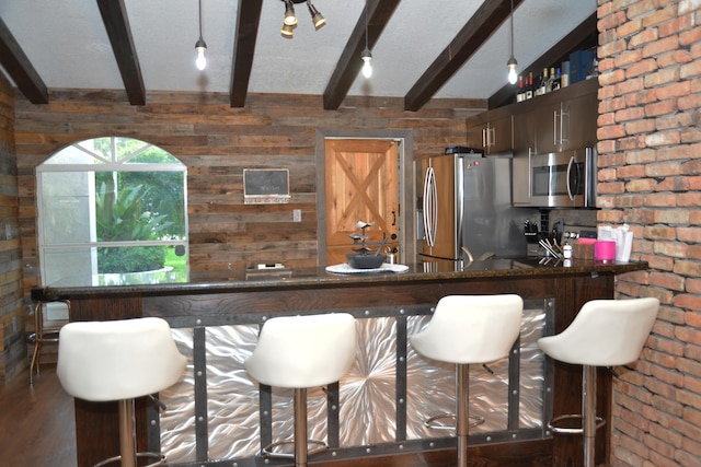 kitchen with dark wood-type flooring, beamed ceiling, appliances with stainless steel finishes, wooden walls, and a breakfast bar area