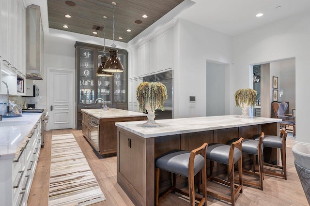 kitchen featuring pendant lighting, a center island with sink, stainless steel built in refrigerator, light wood-type flooring, and light stone counters