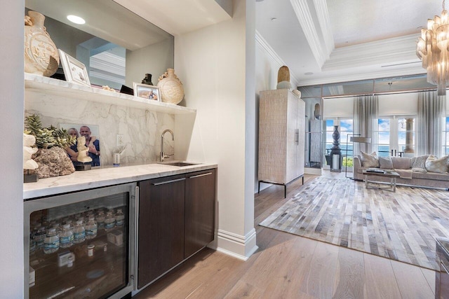 bar with sink, dark brown cabinets, a tray ceiling, beverage cooler, and light wood-type flooring