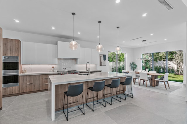 kitchen with an island with sink, pendant lighting, light tile flooring, white cabinetry, and sink