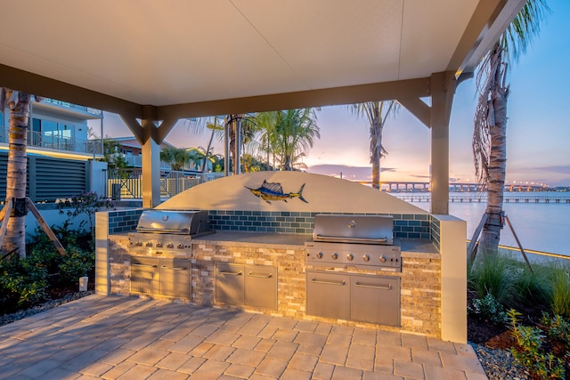 patio terrace at dusk featuring area for grilling, a water view, and an outdoor kitchen