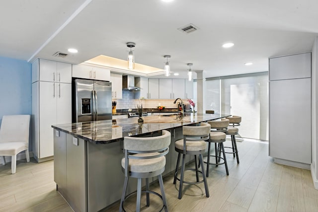 kitchen with a large island with sink, wall chimney exhaust hood, decorative light fixtures, and stainless steel refrigerator with ice dispenser