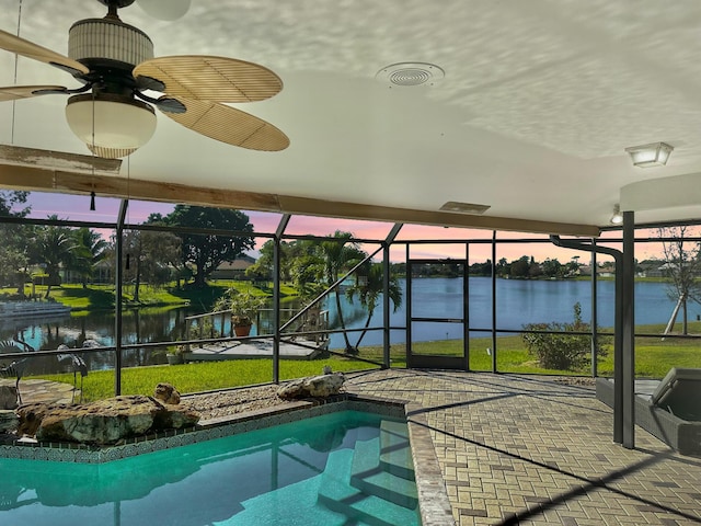 pool at dusk featuring a lanai, ceiling fan, a patio area, and a water view