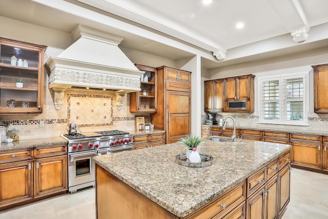 kitchen featuring stainless steel appliances, light stone counters, backsplash, a kitchen island with sink, and custom range hood