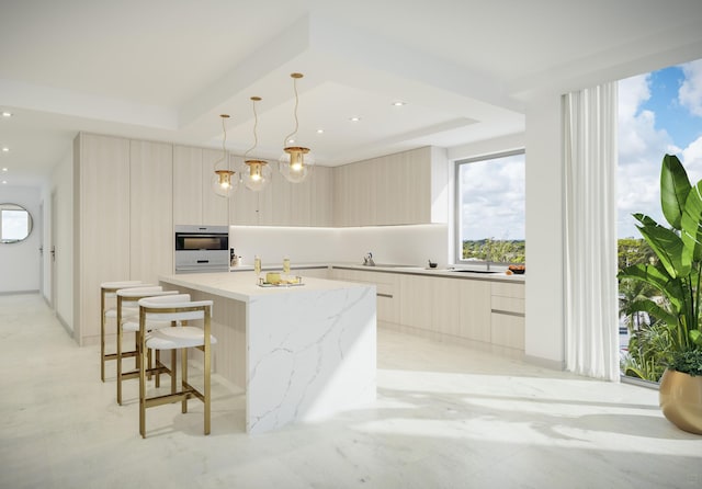 kitchen featuring a tray ceiling, a kitchen island with sink, light brown cabinetry, hanging light fixtures, and light stone countertops