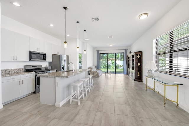 kitchen featuring light stone countertops, decorative light fixtures, white cabinetry, appliances with stainless steel finishes, and a center island with sink