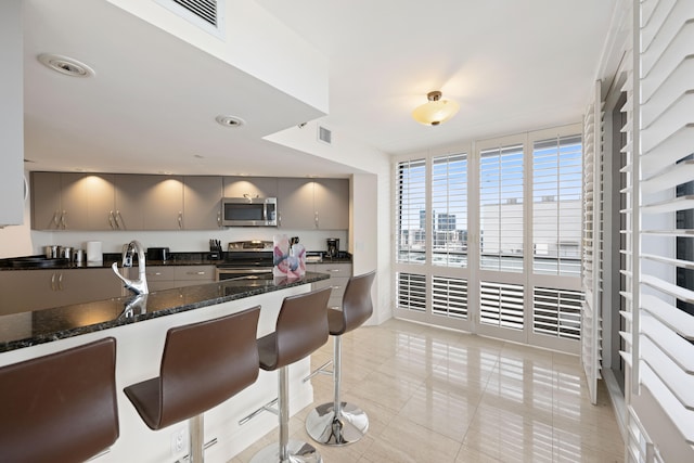 kitchen with gray cabinets, stainless steel appliances, dark stone countertops, a kitchen breakfast bar, and light tile floors