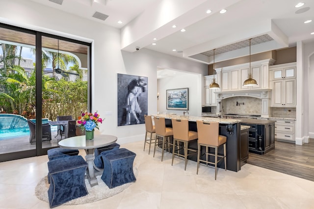 kitchen with light tile floors, backsplash, decorative light fixtures, a kitchen island with sink, and light stone countertops