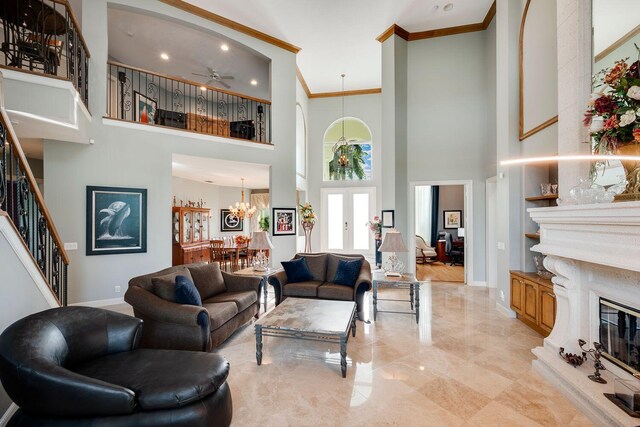 living room featuring french doors, crown molding, a towering ceiling, a fireplace, and ceiling fan with notable chandelier