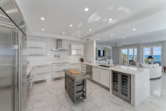 kitchen with sink, wall chimney exhaust hood, kitchen peninsula, white cabinets, and appliances with stainless steel finishes